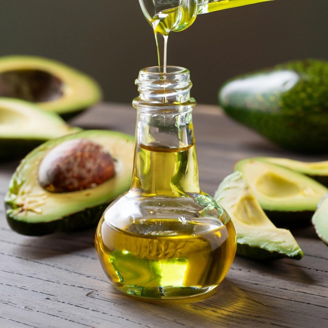 Image of avocado oil being poured into small ornate glass jars with avocados scattered on the table..